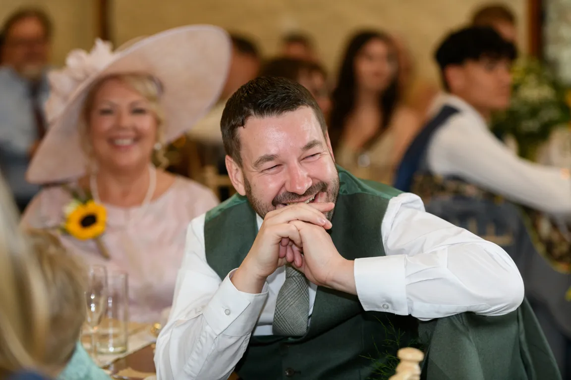 Smiling man in white shirt and green vest seated at a table with a woman wearing a large hat and a sunflower corsage in the background at a formal event.