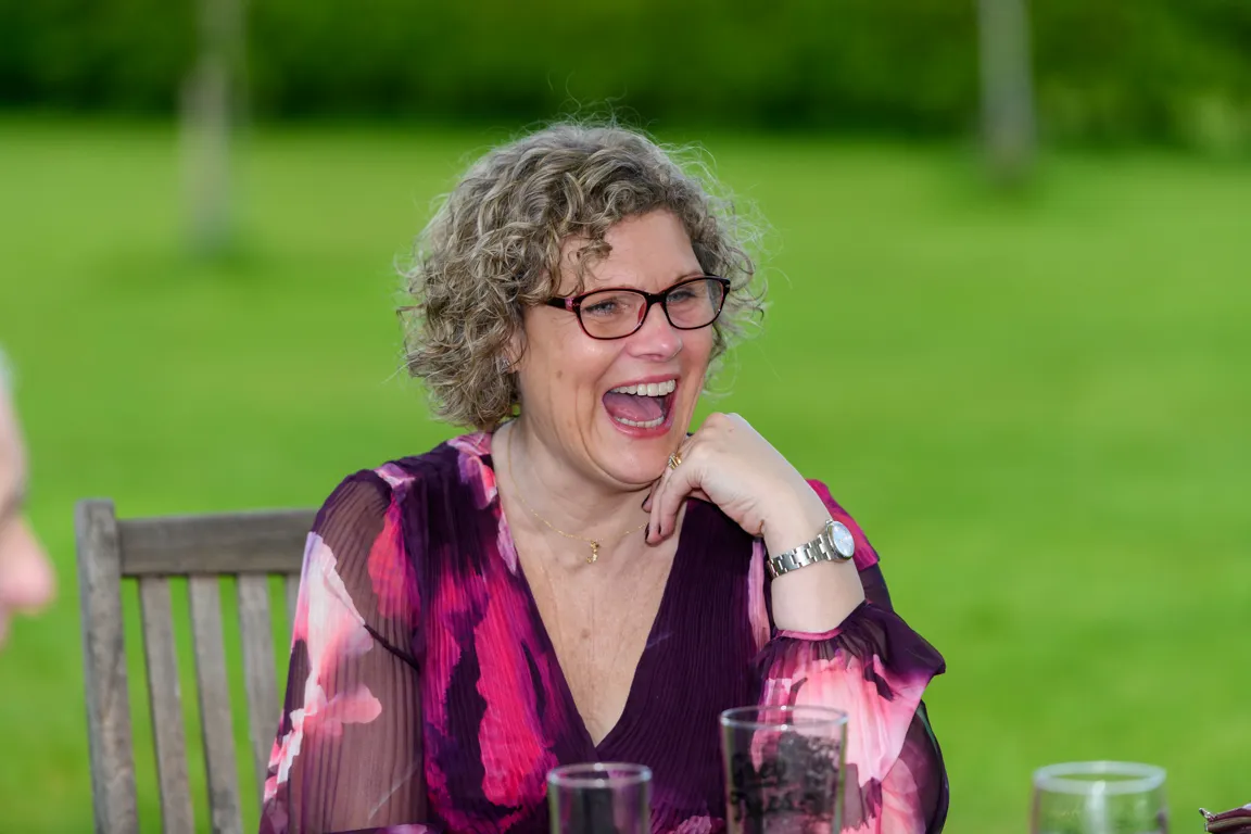 Woman with curly hair and glasses laughing while sitting outdoors at a table with drinks.