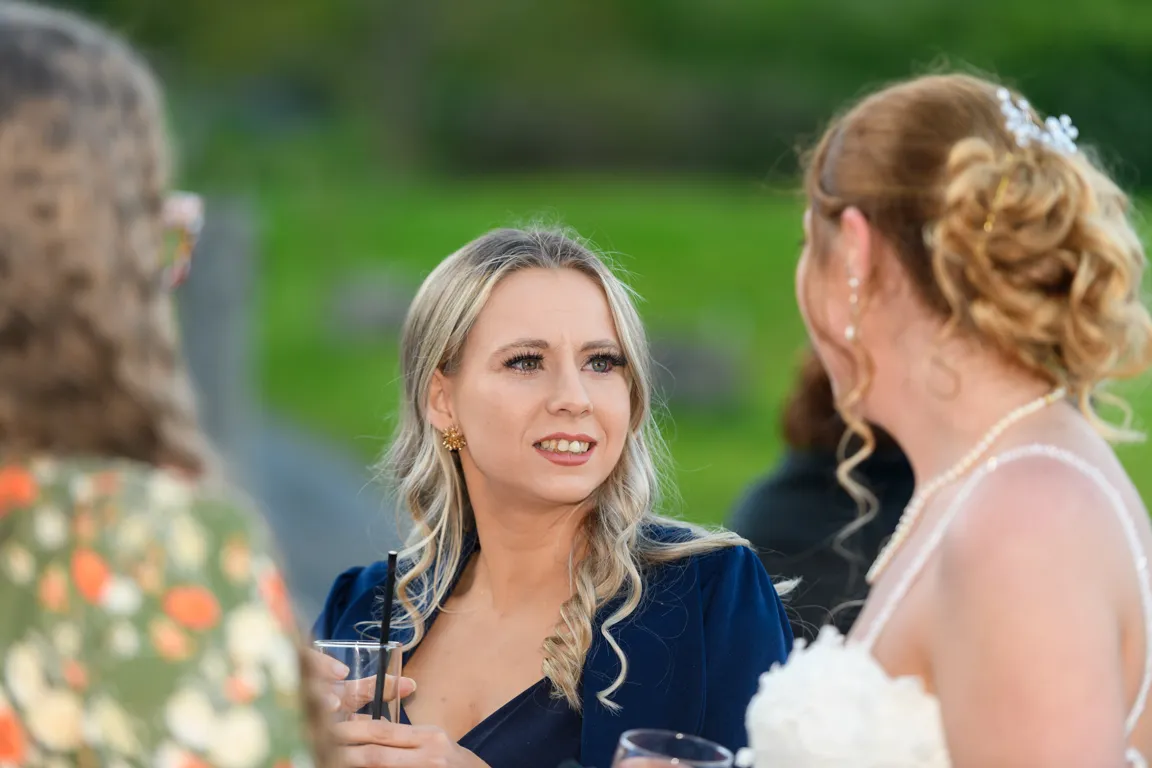Blonde woman in a navy dress holding a drink and talking to a bride in a white wedding dress with pearl necklace and hair accessory.