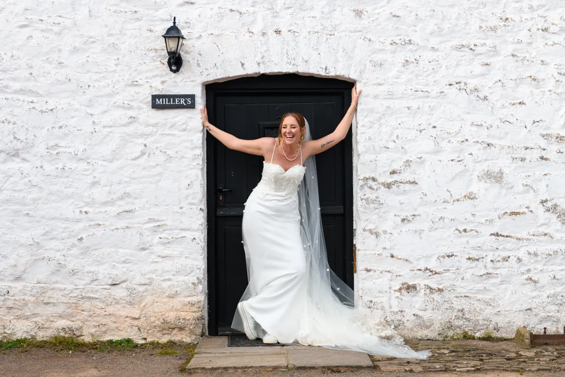 Bride in a white wedding dress and veil laughing while standing in a small black doorway set in a white stone wall.