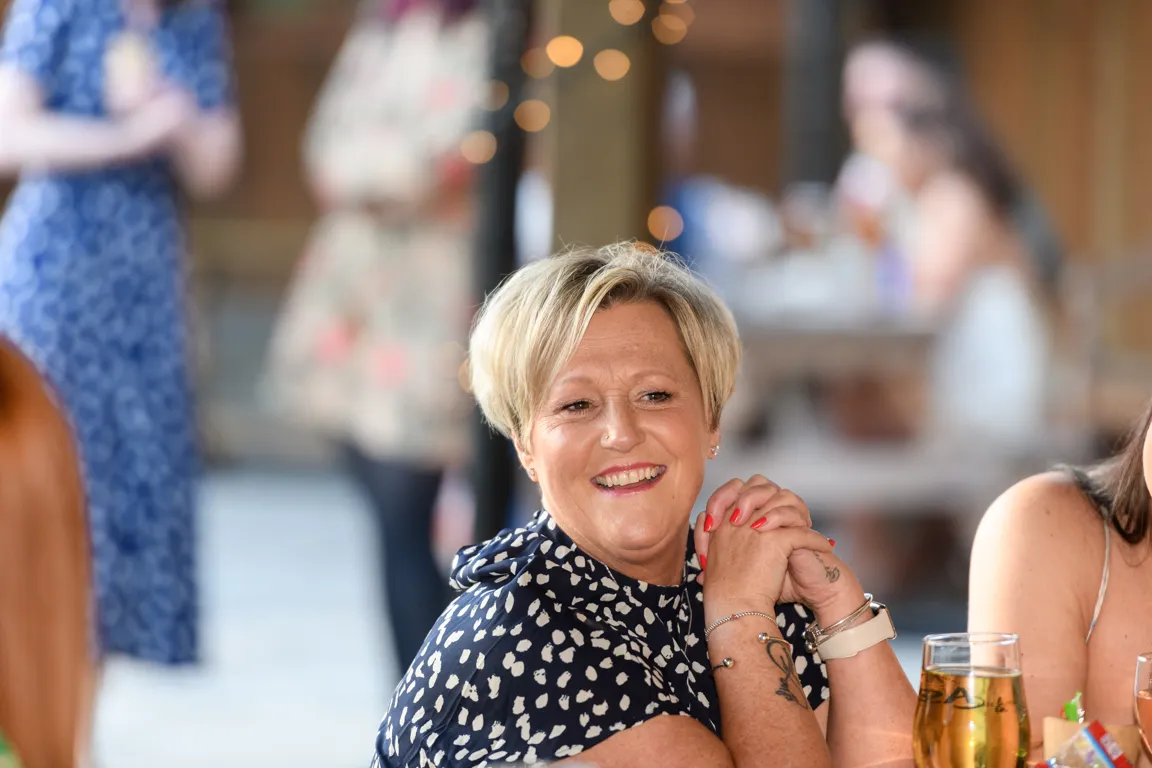 Smiling woman with short blonde hair wearing a navy blue and white patterned blouse, seated at a table with drinks, hands clasped.