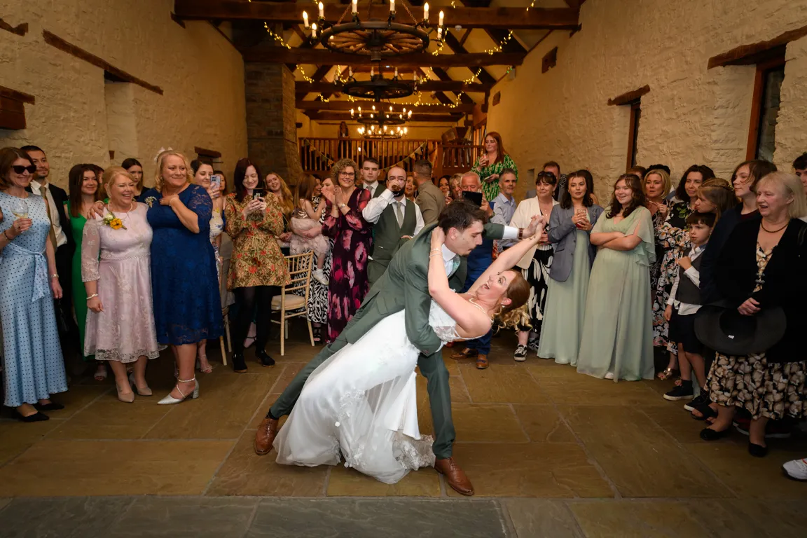 Bride and groom in a deep dance dip surrounded by guests in a rustic stone-walled venue with chandeliers.