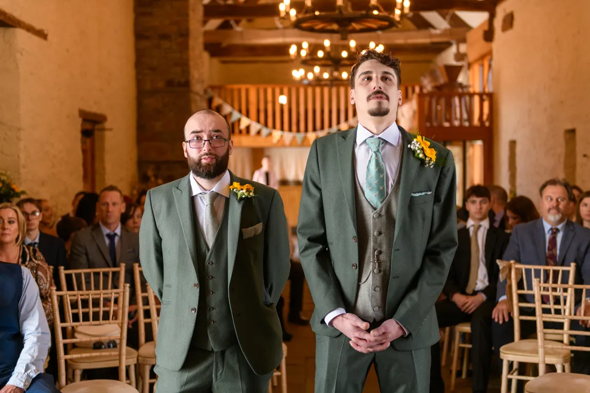 Two groomsmen in green suits with yellow boutonnieres standing at a wedding ceremony with guests seated behind them.
