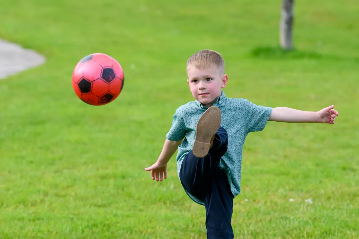 Young boy in a green patterned shirt and dark pants kicking a red soccer ball on a grassy field.