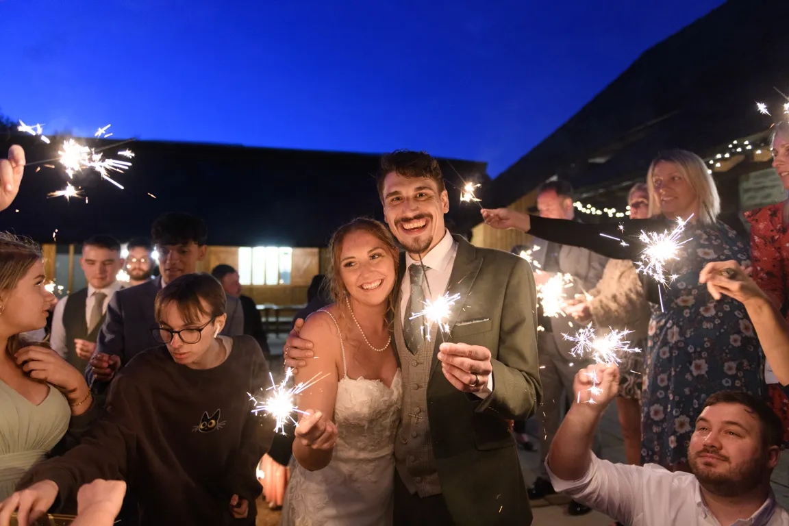 A smiling bride in a white dress and groom in a suit holding sparklers surrounded by guests holding sparklers at an outdoor evening celebration.
