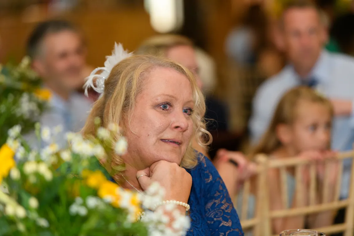 Woman in blue lace dress with a feather hair accessory resting her chin on her hand, surrounded by blurred people and flowers indoors.