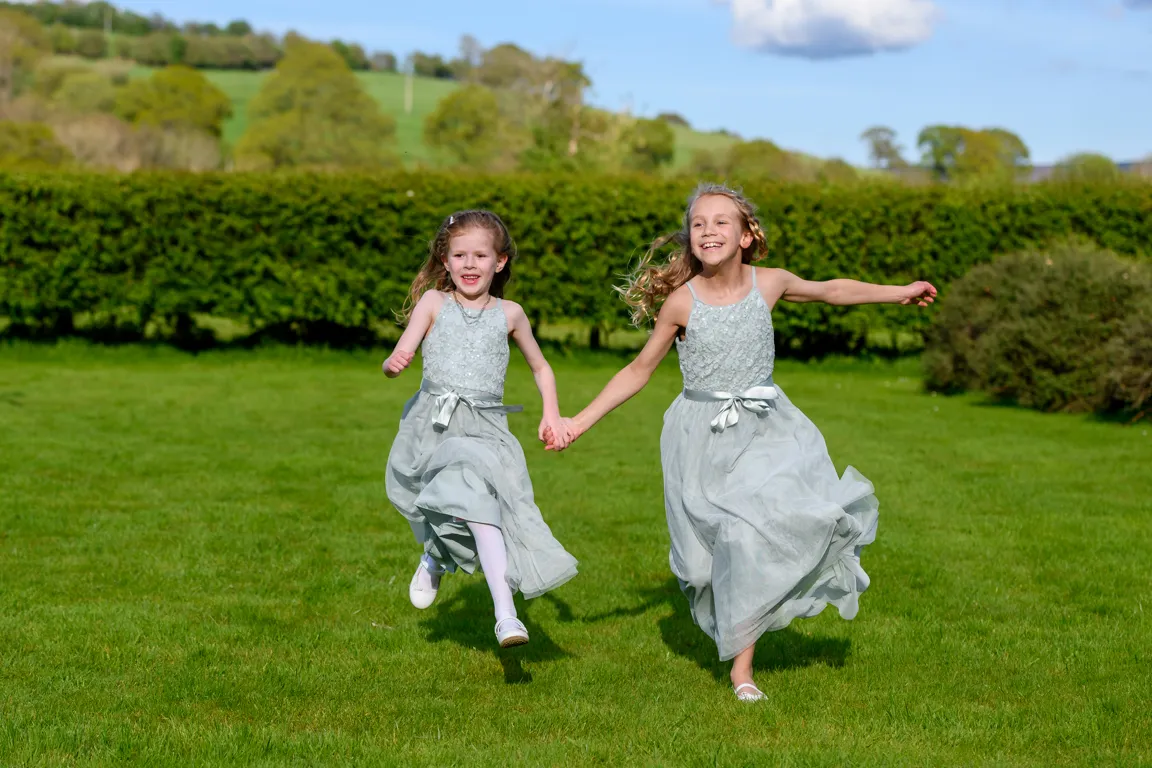Two young girls in matching light blue dresses running hand in hand on green grass with trees in the background.