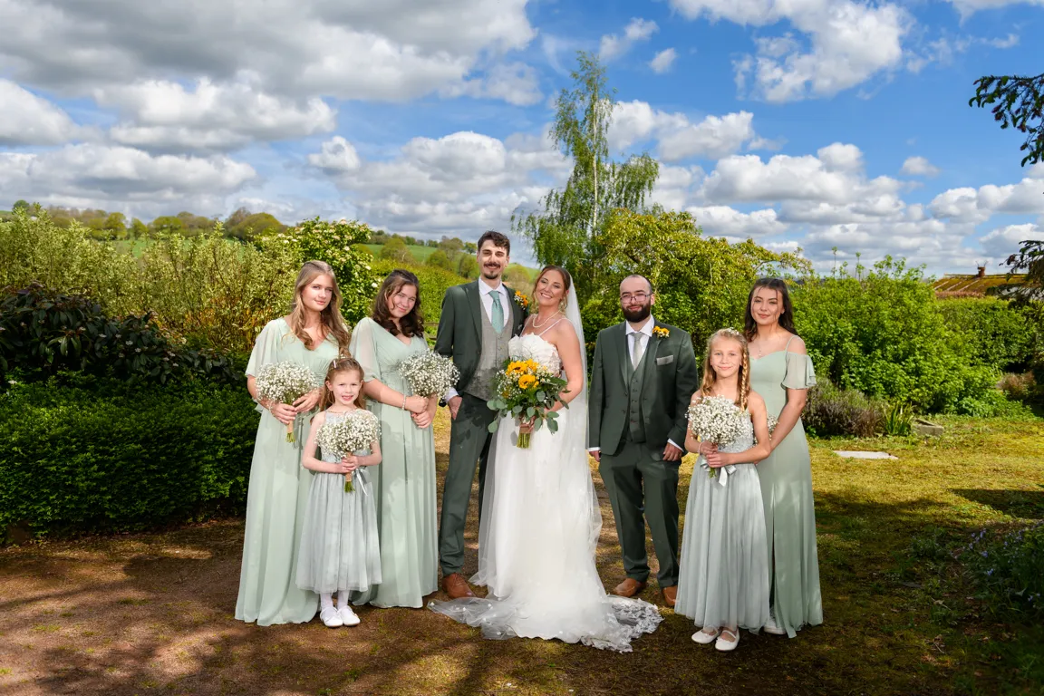 Bride and groom with bridesmaids and flower girls dressed in light green attire standing outdoors with blue sky and green foliage background.