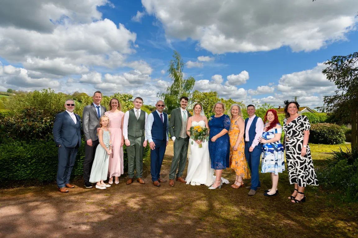 Group of twelve people posing outdoors on a sunny day with cloudy blue sky, including a bride in a white dress holding a bouquet and a groom in a green suit.
