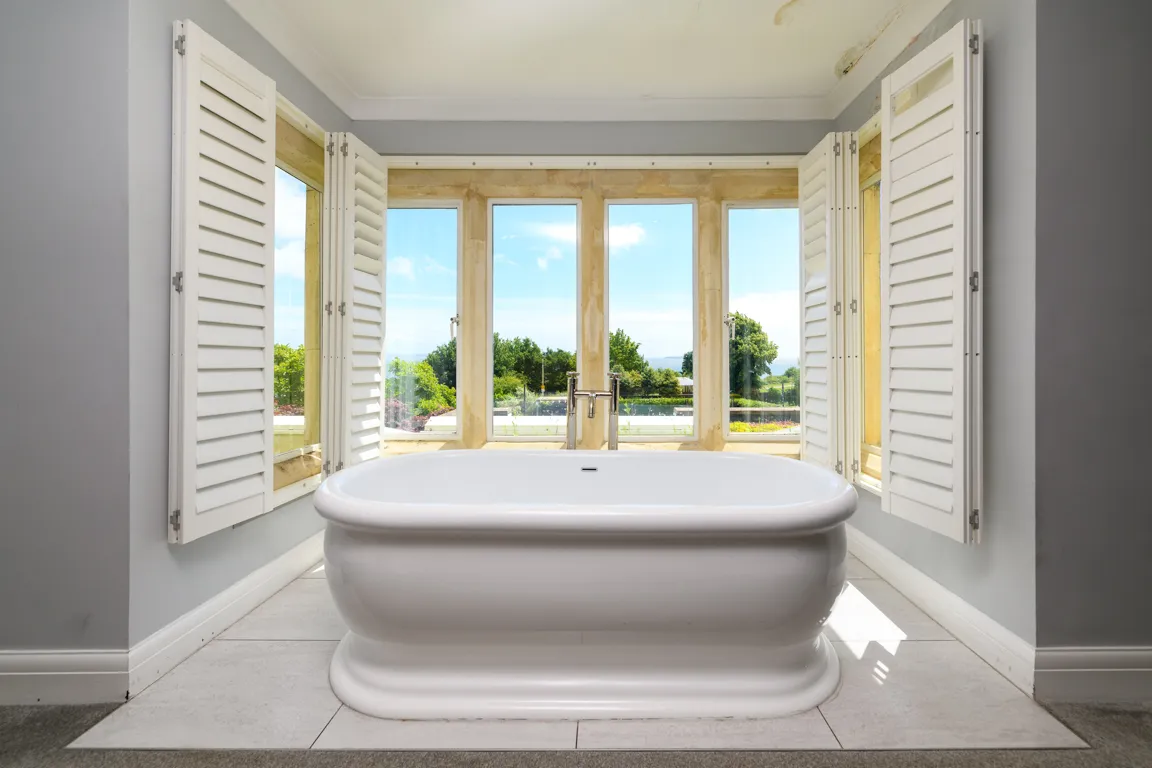 White freestanding bathtub centered in front of large windows with open white shutters, overlooking greenery outside.