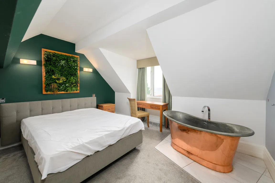 Modern bedroom with slanted ceilings featuring a gray upholstered bed, copper bathtub, wooden desk, green accent wall, and vertical garden.