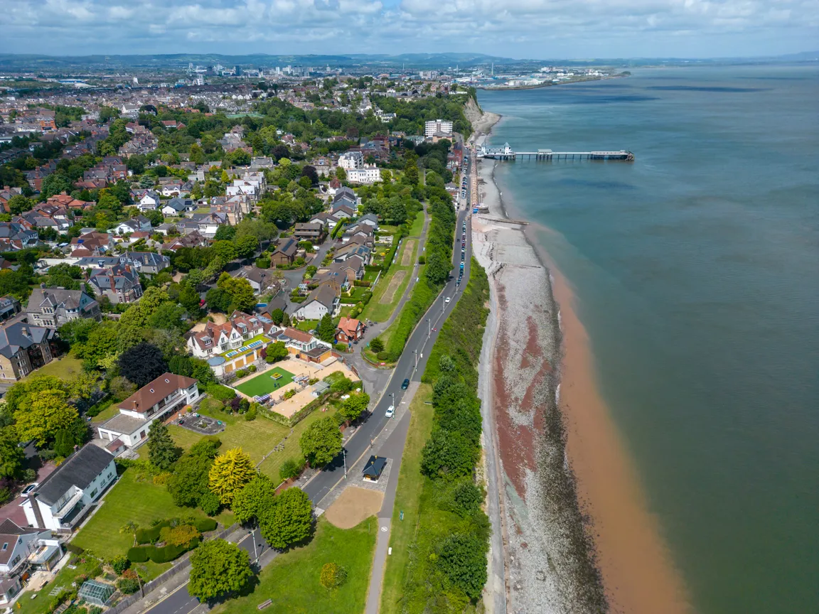 Aerial view of a coastal town with residential houses, a road along the shoreline, green trees, and a pier extending into the sea under a partly cloudy sky.