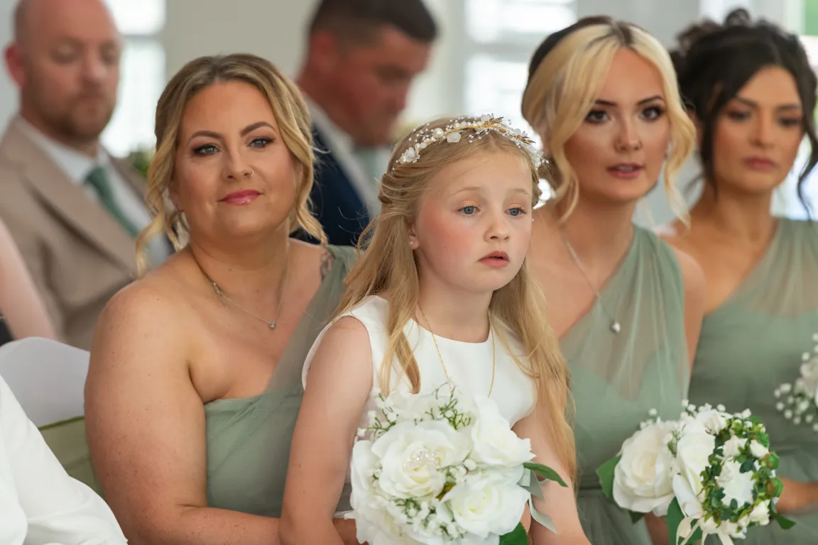 Four women and a young girl wearing green bridesmaid dresses and holding white flower bouquets at a wedding ceremony.
