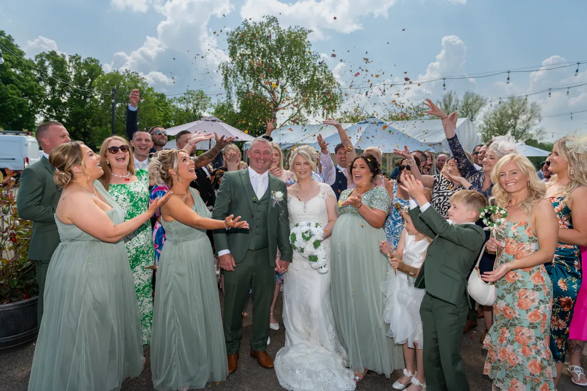 Newlywed bride in white lace dress and groom in green suit smiling surrounded by wedding guests throwing confetti outdoors.