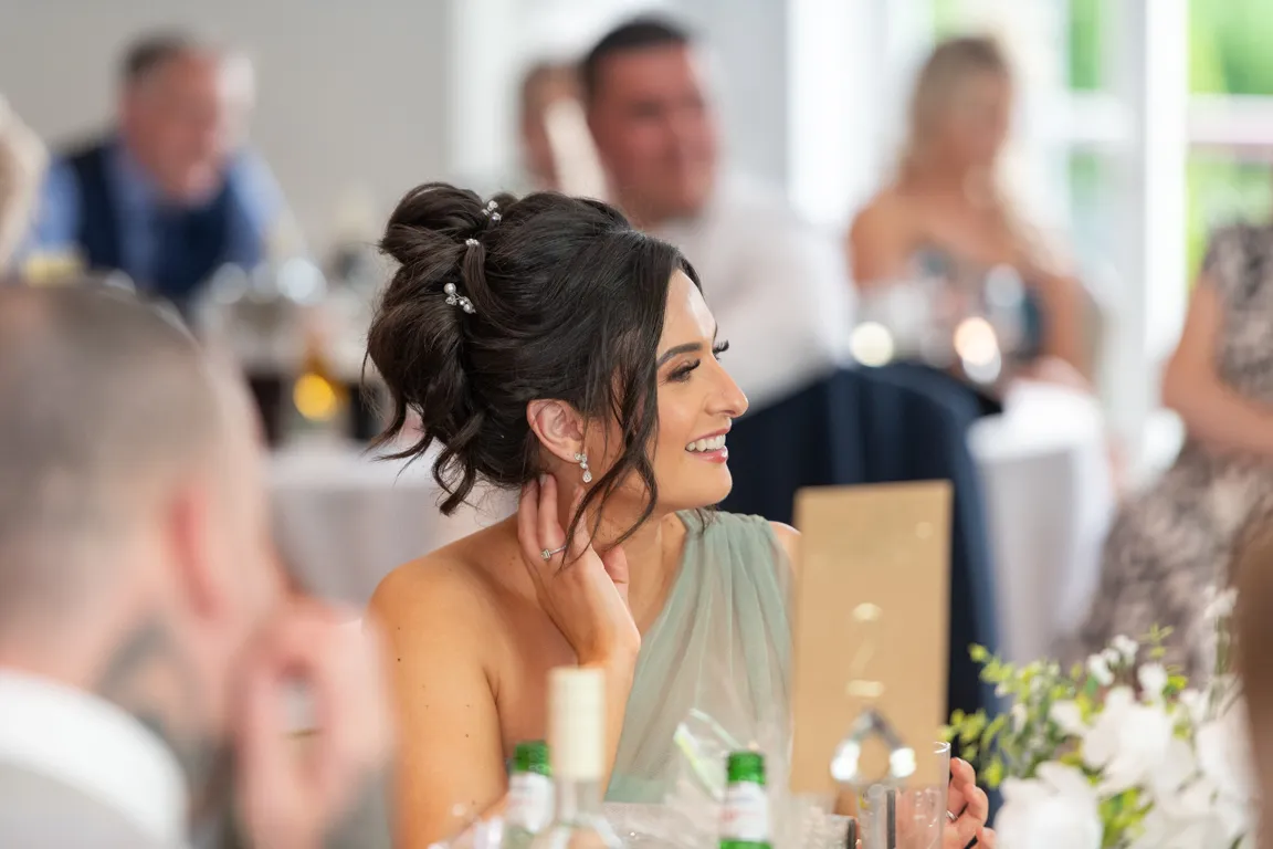 Woman with dark styled hair and earrings smiling at a formal event with blurred guests in background.