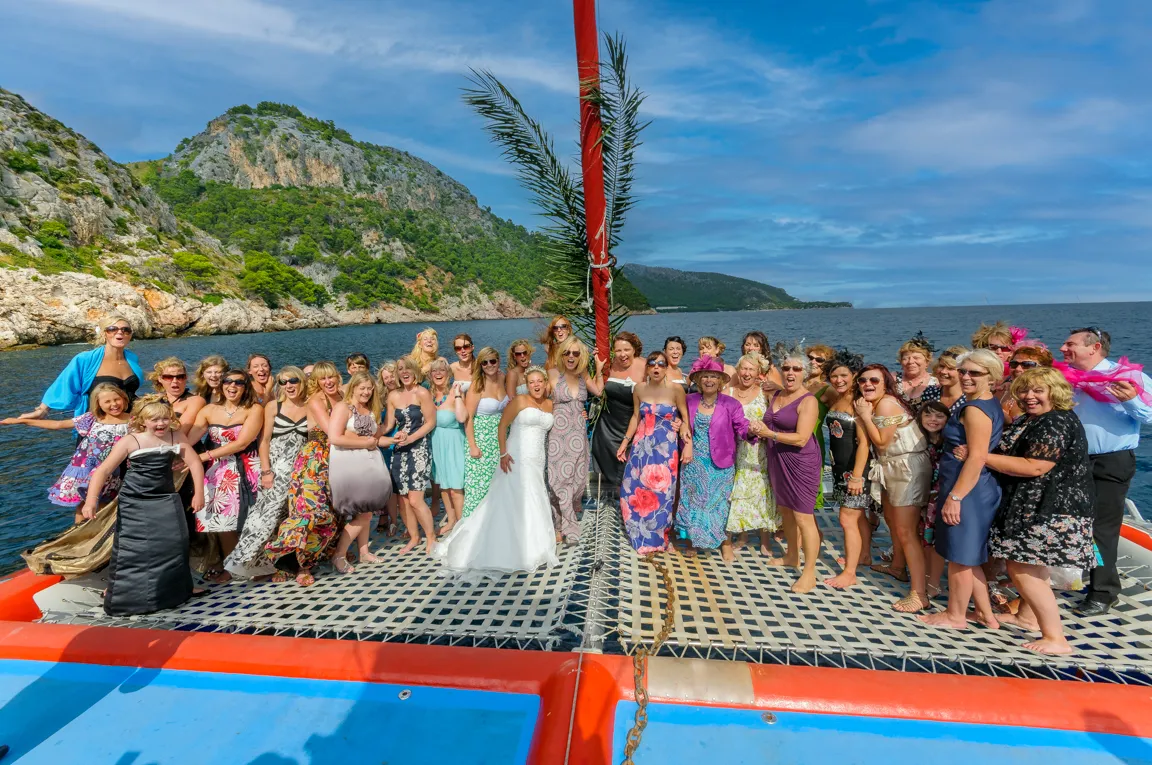 Large group of women dressed in suits, dresses, and a bride in a wedding gown posing on a boat net with rocky hills and ocean in the background.