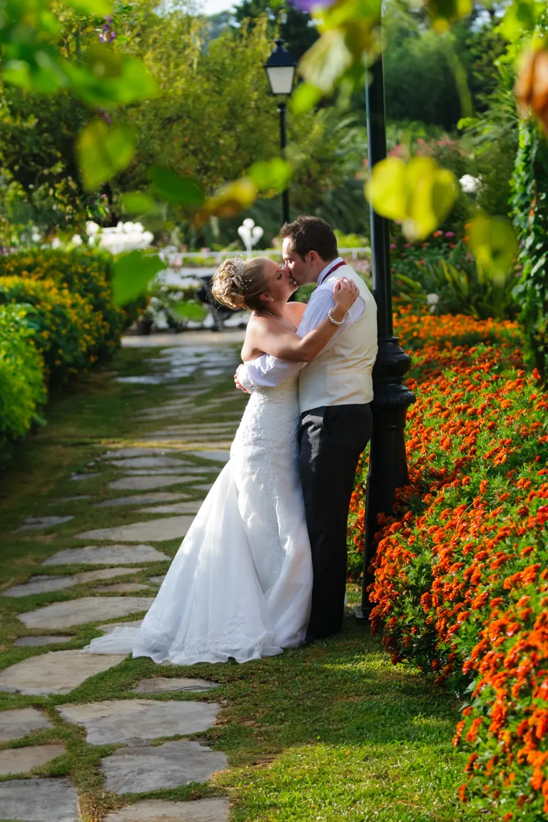 Bride and groom embracing and kissing in a sunny garden with stone path and vibrant orange flowers.