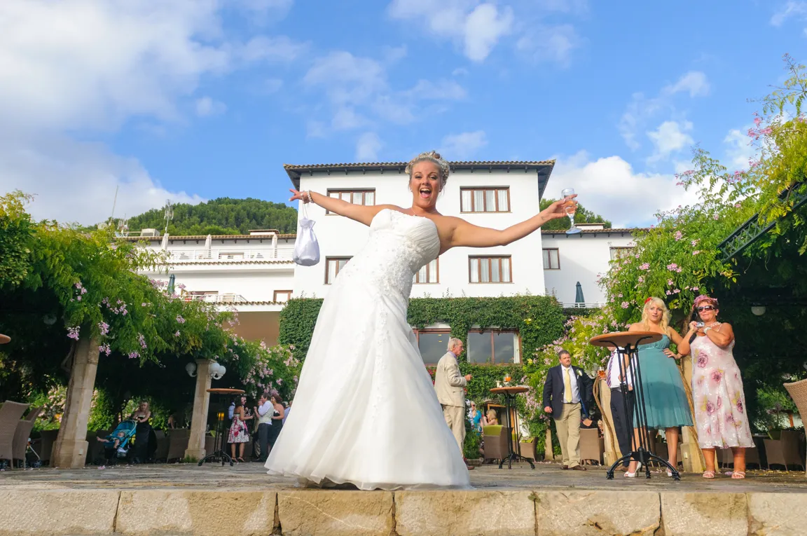 Joyful bride in a white wedding dress holding a glass of wine and a bag, with guests and a white building with greenery in the background.