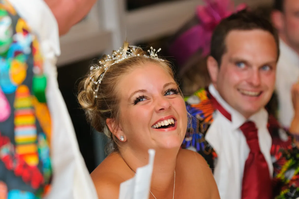 Smiling bride with a jeweled tiara looking up during a wedding celebration, with a man in a colorful jacket and red tie in the background.