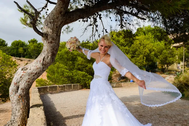 Smiling bride in a white wedding gown holding a bouquet and posing outdoors near a tree with greenery in the background.
