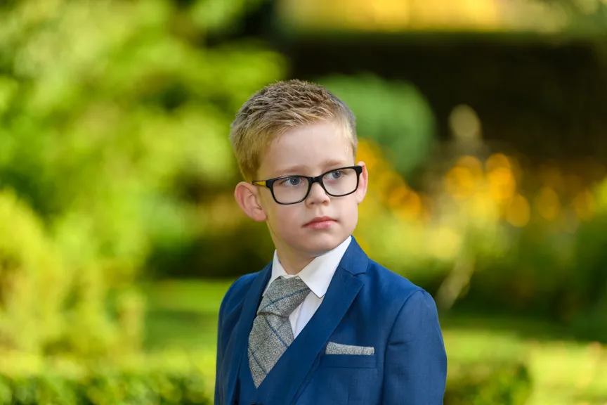 Young boy in glasses wearing a blue suit and patterned tie standing outdoors with a blurred green and yellow background.