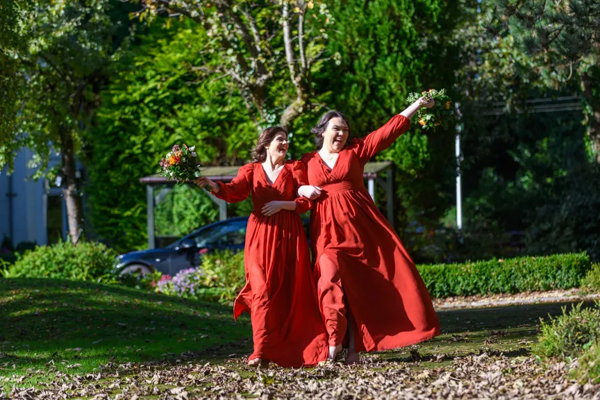 Two women in matching red dresses joyfully walking arm in arm outdoors, each holding a bouquet.