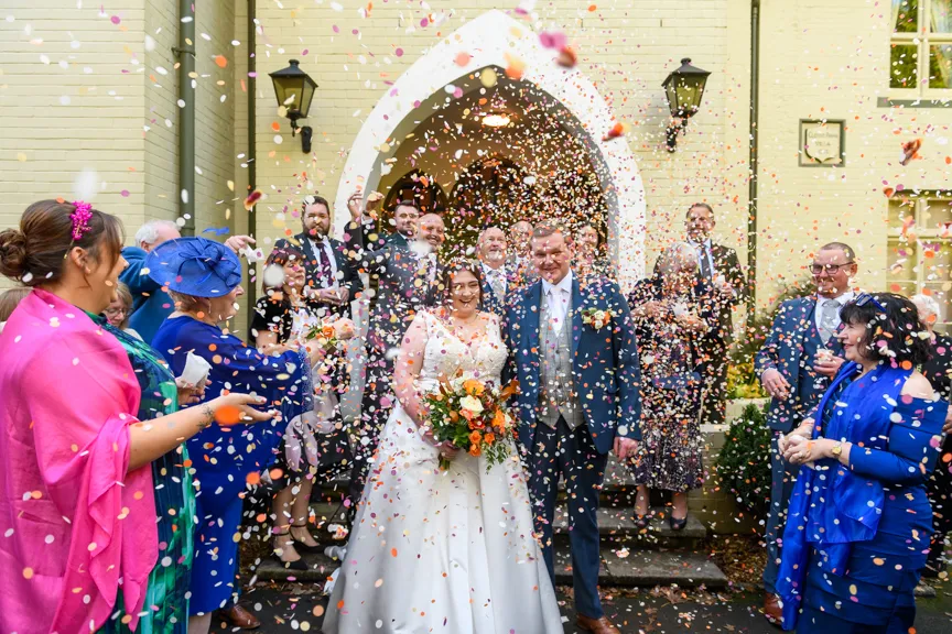 Bride and groom standing outside a building doorway smiling, surrounded by guests throwing colorful confetti.