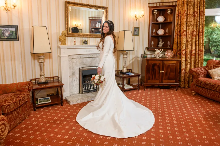 Bride in a long white wedding dress holding a bouquet, standing in a vintage-style living room with patterned red carpet and furniture.