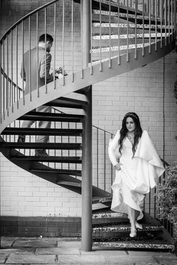 Bride in wedding dress descending a spiral staircase with groom holding bouquet standing above her.