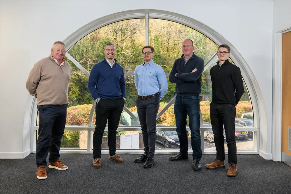 Five men standing indoors in front of a large semicircular window with a view of trees and parked cars outside.