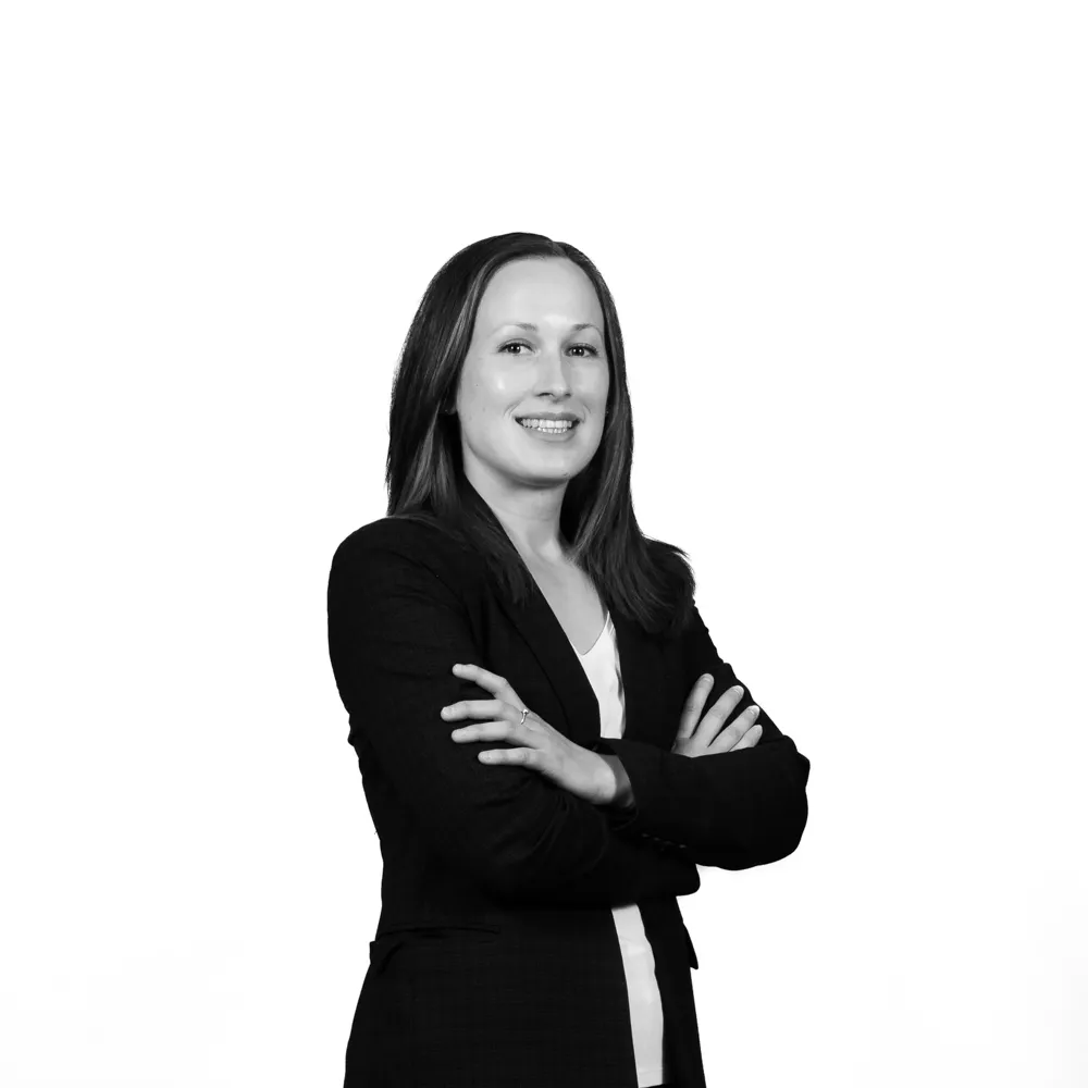 Confident woman with arms crossed wearing a dark blazer and white blouse, smiling at the camera on a white background.