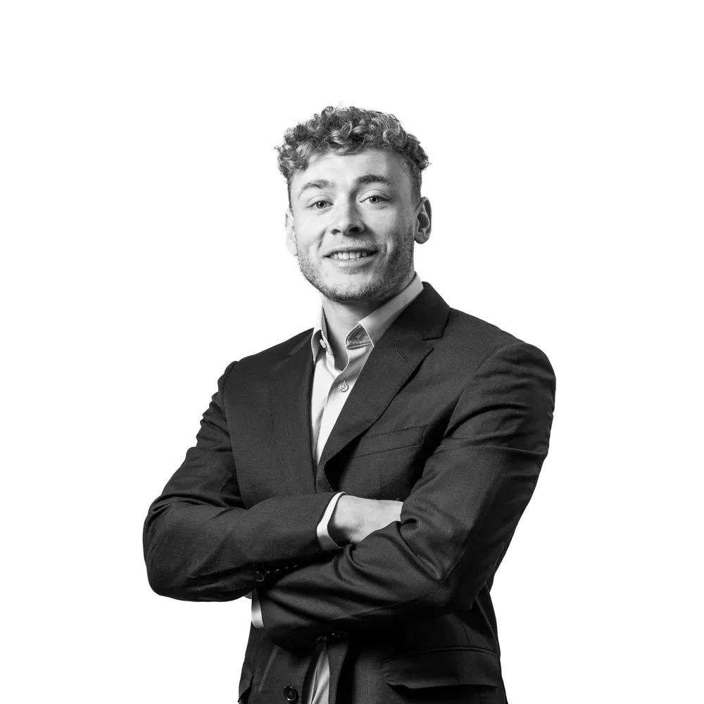 Smiling young man with curly hair wearing a dark suit jacket and light shirt crossing his arms.