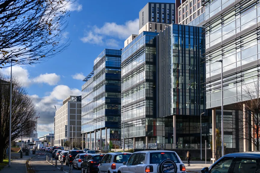 Busy urban street with modern glass office buildings on the right and a line of cars in traffic under a partly cloudy blue sky.