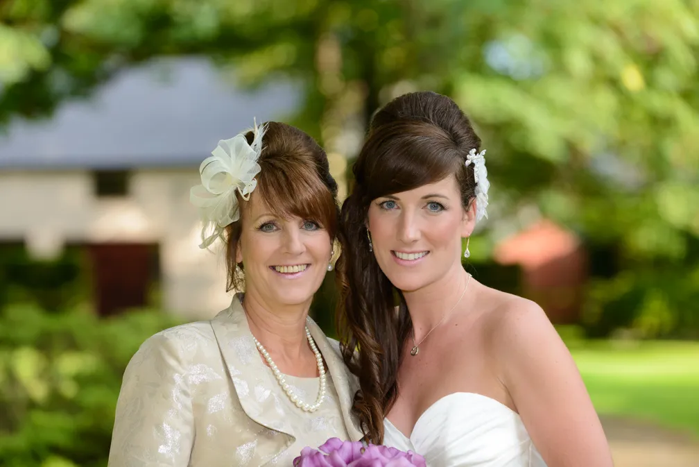 Two women smiling outdoors; one in a white wedding dress holding a bouquet of purple flowers, the other in a beige suit with a pearl necklace and decorative hairpiece.