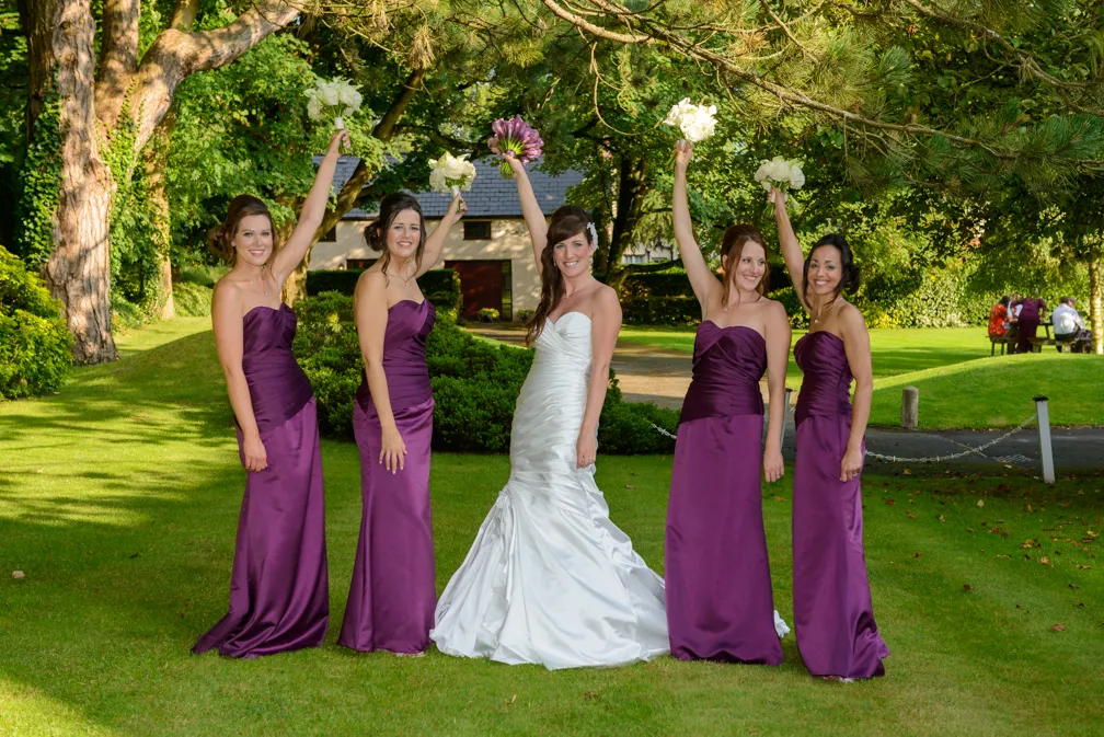 Bride in a white gown standing on green lawn with four bridesmaids in purple dresses holding bouquets up.