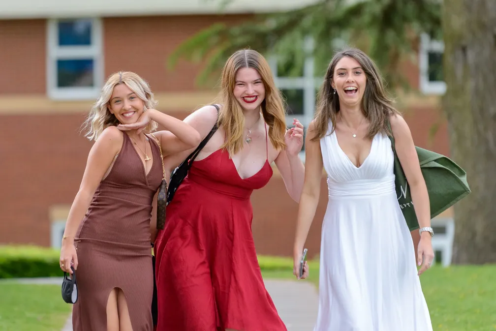 Three young women smiling and walking outdoors, wearing dresses in brown, red, and white.