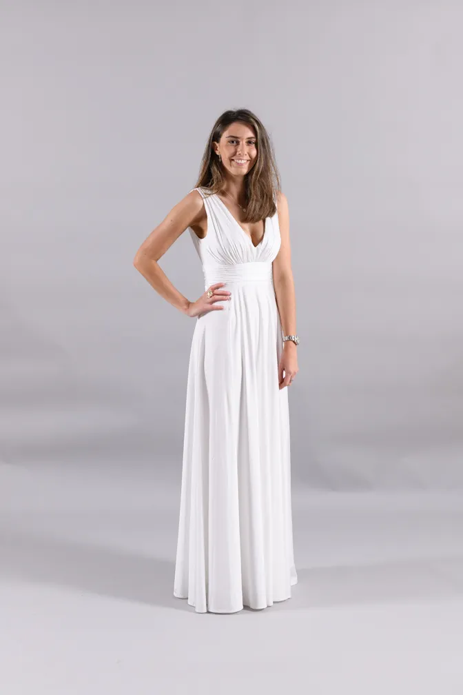 Woman smiling wearing a long white sleeveless dress standing against a plain light gray background.