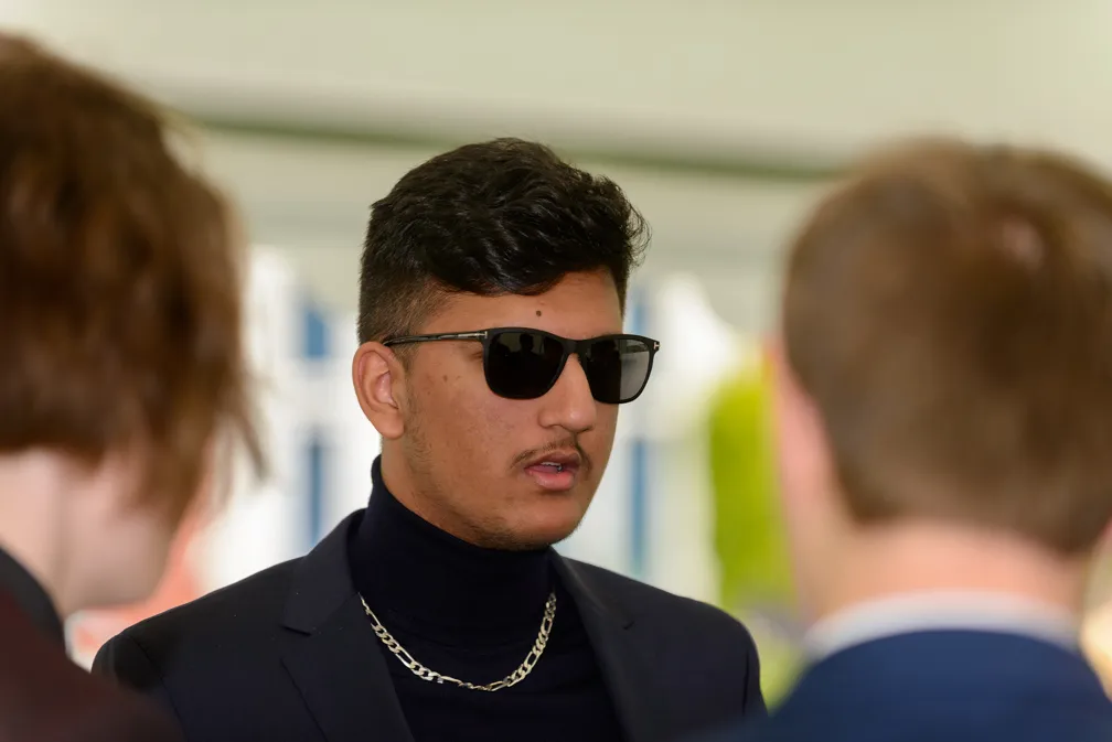 Young man wearing a dark blazer, black turtleneck, silver chain, and sunglasses, speaking with two other people.
