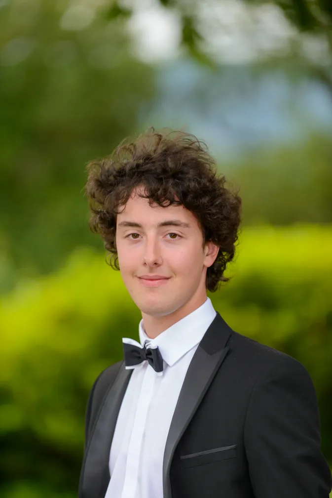 Young man with curly hair wearing a black tuxedo and bow tie, smiling with blurred greenery background.
