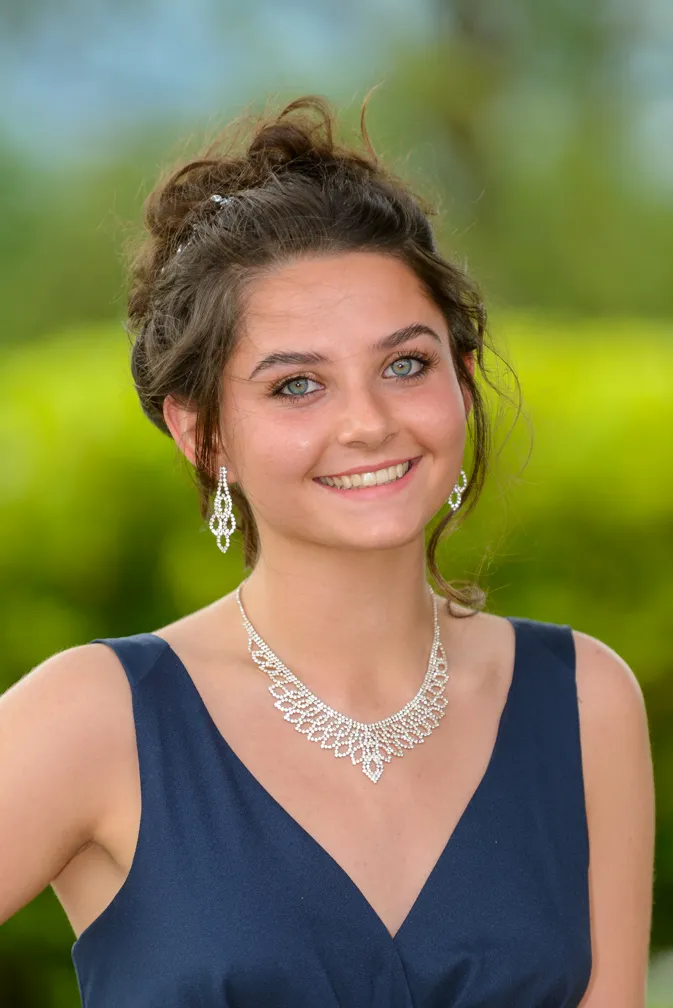 Young woman with green eyes and curly hair in an updo wearing a dark blue sleeveless dress and silver jewelry including earrings and a necklace, smiling against a blurred green outdoor background.