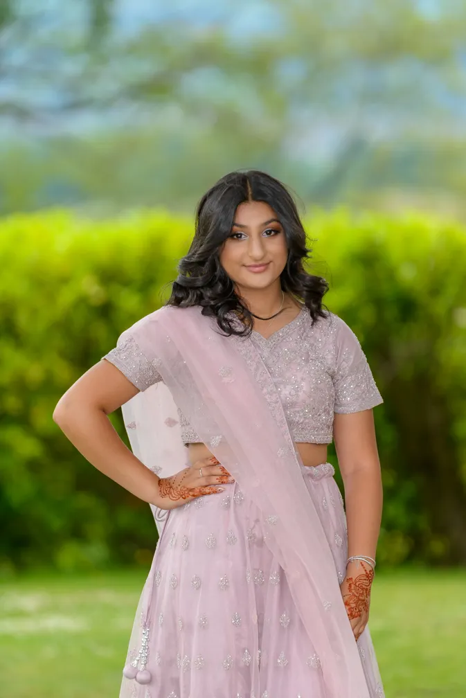 Young woman wearing a light pink traditional dress with henna on her hands, standing outdoors with greenery blurred in the background.