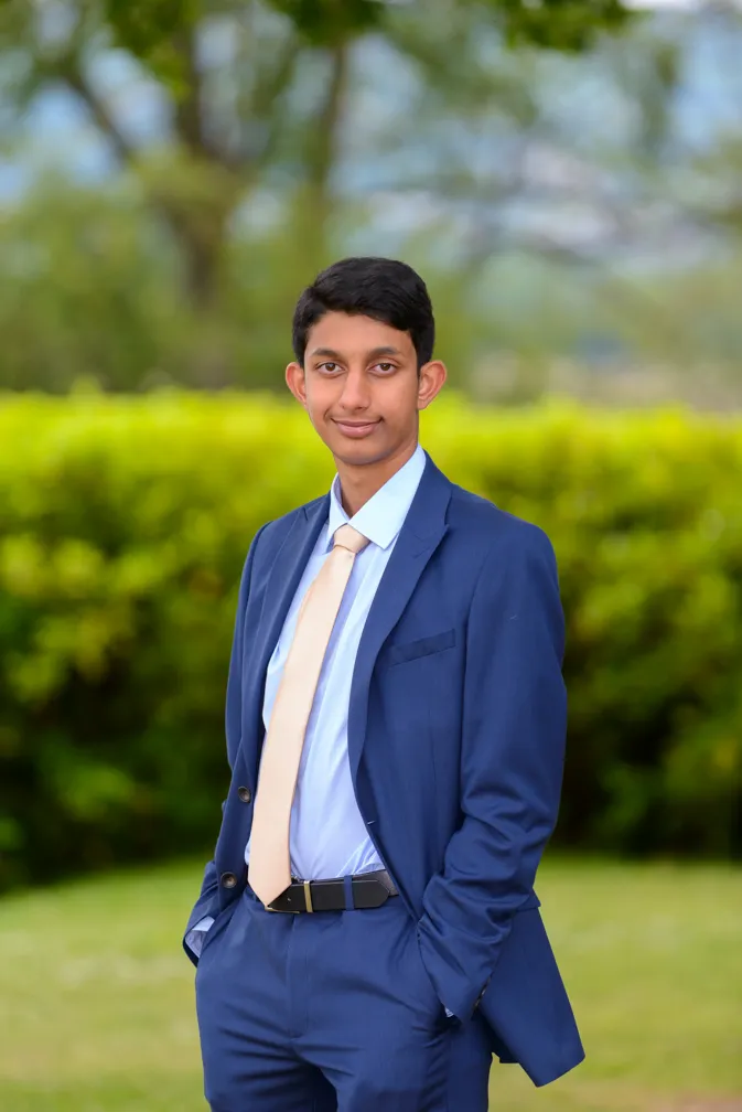 Young man in a blue suit with a light beige tie standing outdoors with hands in pockets against a blurred green hedge background.
