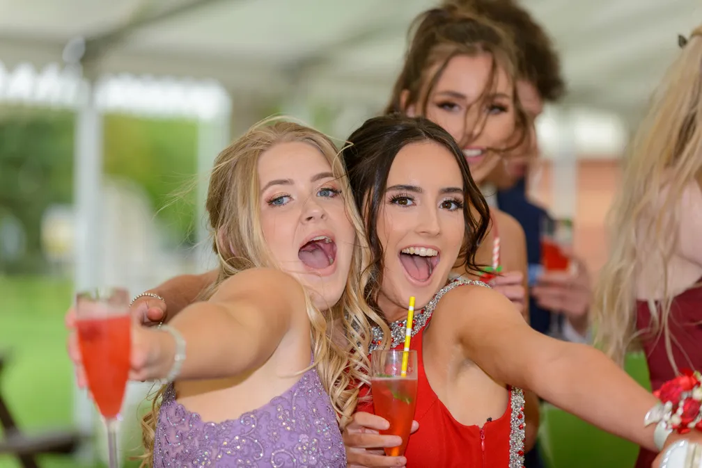 Two young women smiling excitedly while holding red drinks at an outdoor event under a tent.