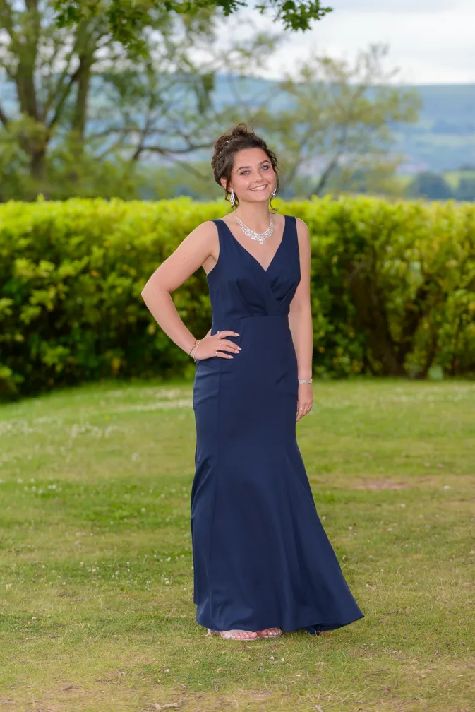 Smiling young woman in a navy blue sleeveless evening gown standing on grass with greenery and hills in the background.