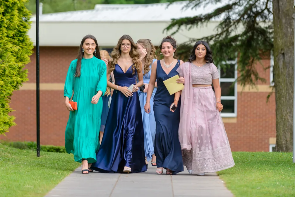 Group of five young women dressed in elegant formal gowns walking outdoors on a paved path with greenery and a building in the background.
