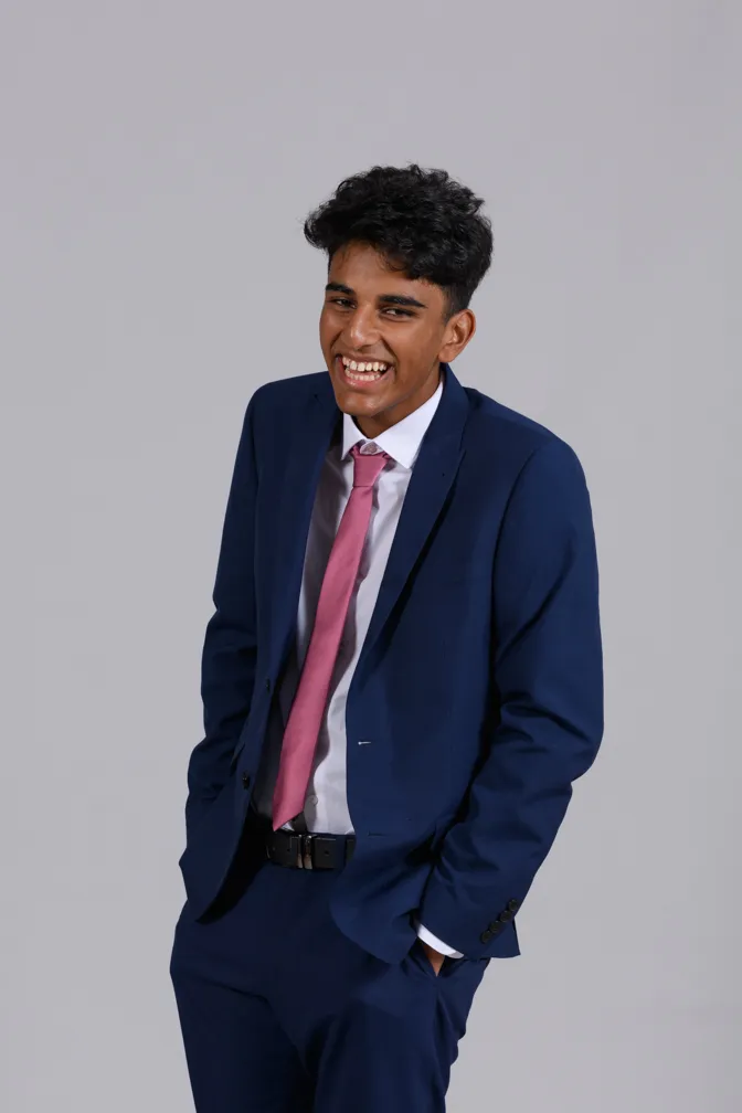 Young man in a blue suit with a pink tie smiling with hands in pockets against a plain gray background.