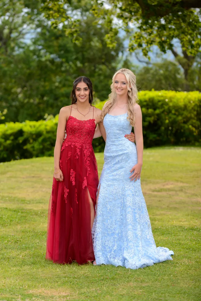 Two young women wearing elegant long dresses, one in red and one in light blue, posing together outdoors on grass with trees in the background.