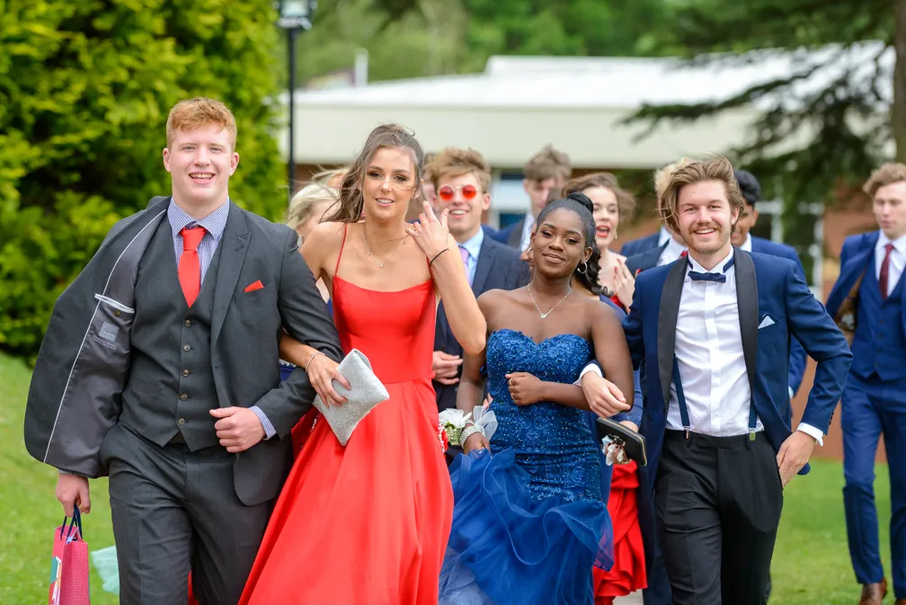 Group of young people dressed in formal attire walking outside on grass, with men in suits and women in vibrant red and blue gowns.