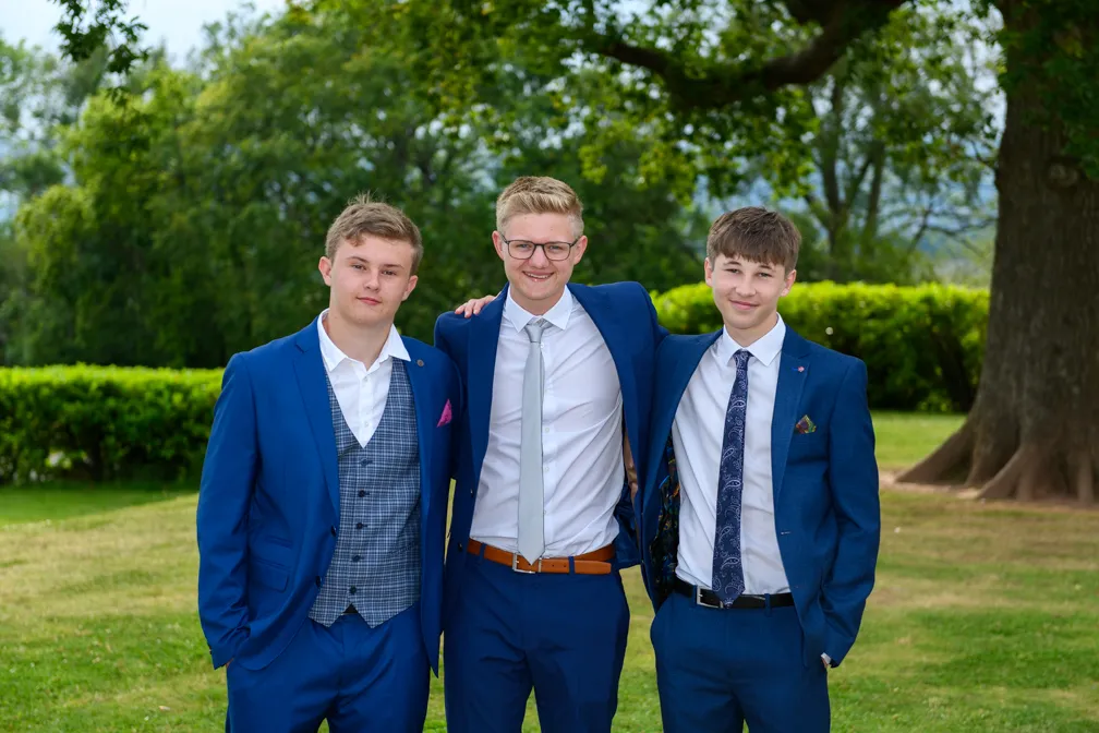 Three teenage boys in blue suits standing outdoors on grass with trees in the background, smiling and posing together.