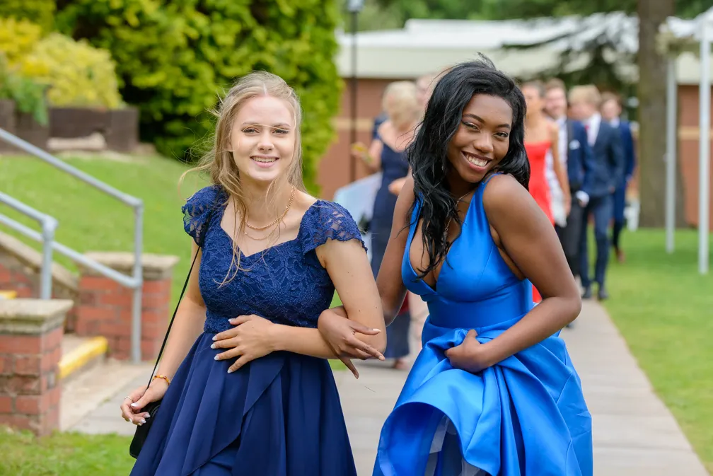 Two young women smiling and walking arm in arm in blue formal dresses at an outdoor event.